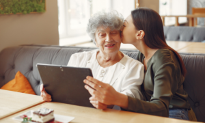 A young woman kisses her grandmother's cheek as they look at a tablet together