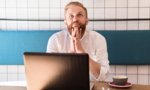 A man in a cafe with a laptop, looking up while thinking