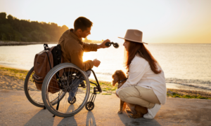 A young man in a wheelchair on a beach with a woman and small dog